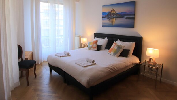 A hotel room with a large bed, two bedside lamps, a painting on the wall, and a chair. - Libération (Nice)