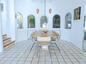 A dining area with a large table, chairs, and a bowl of fruit. There are windows with arched tops and a view of greenery outside.