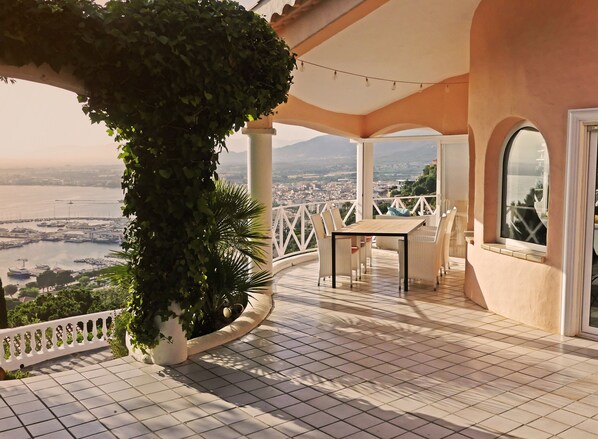 A terrace with a table and chairs, surrounded by white railings and potted plants, overlooking a marina and cityscape.
