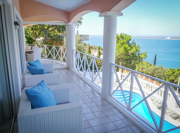 A balcony with white wicker furniture and blue cushions, overlooking a pool and the sea.