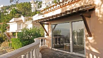 Un balcon doté d’un sol en pierre, d’une balustrade blanche et offrant une vue sur un jardin et des bâtiments en arrière-plan.