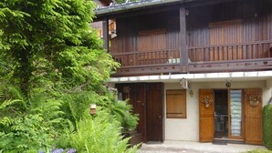 A two-story wooden chalet with a balcony, surrounded by lush greenery and flowering plants. - Cozy 1BR with terrace in Arêches, sleeps 4, near slopes and shuttle (Arêches-Beaufort)