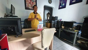 A man in a yellow shirt standing in a room with a desk, computer monitors, and various office items. - OYO 2302 Blue House Costel Syariah (Cianjur)