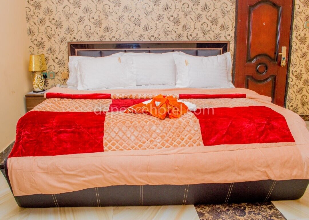Una cama bien tendida con una colcha rojo y beig, un cabecero de madera y un papel tapiz estampado.