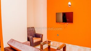 A modern living room with a sofa, armchair, and glass coffee table. A flat-screen TV is mounted on the orange wall. - Peace Hotel and Suites (Ilorin)