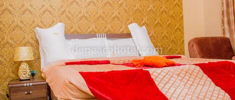 Una cama bien hecha con una colcha roja y color beige, una mesita de noche de madera con una lámpara y un papel tapiz estampado.