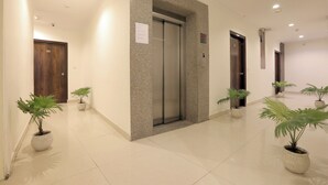 A clean, modern hallway with an elevator, potted plants, and a wooden door. - Castle Blue By Excellent Hospitality (New Delhi)