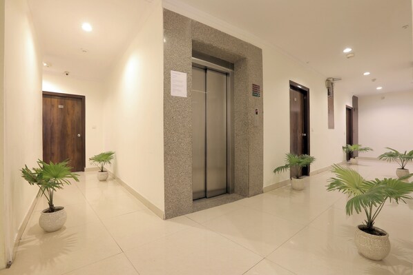 A clean, modern hallway with an elevator, potted plants, and a wooden door. - Castle Blue By Excellent Hospitality (New Delhi)