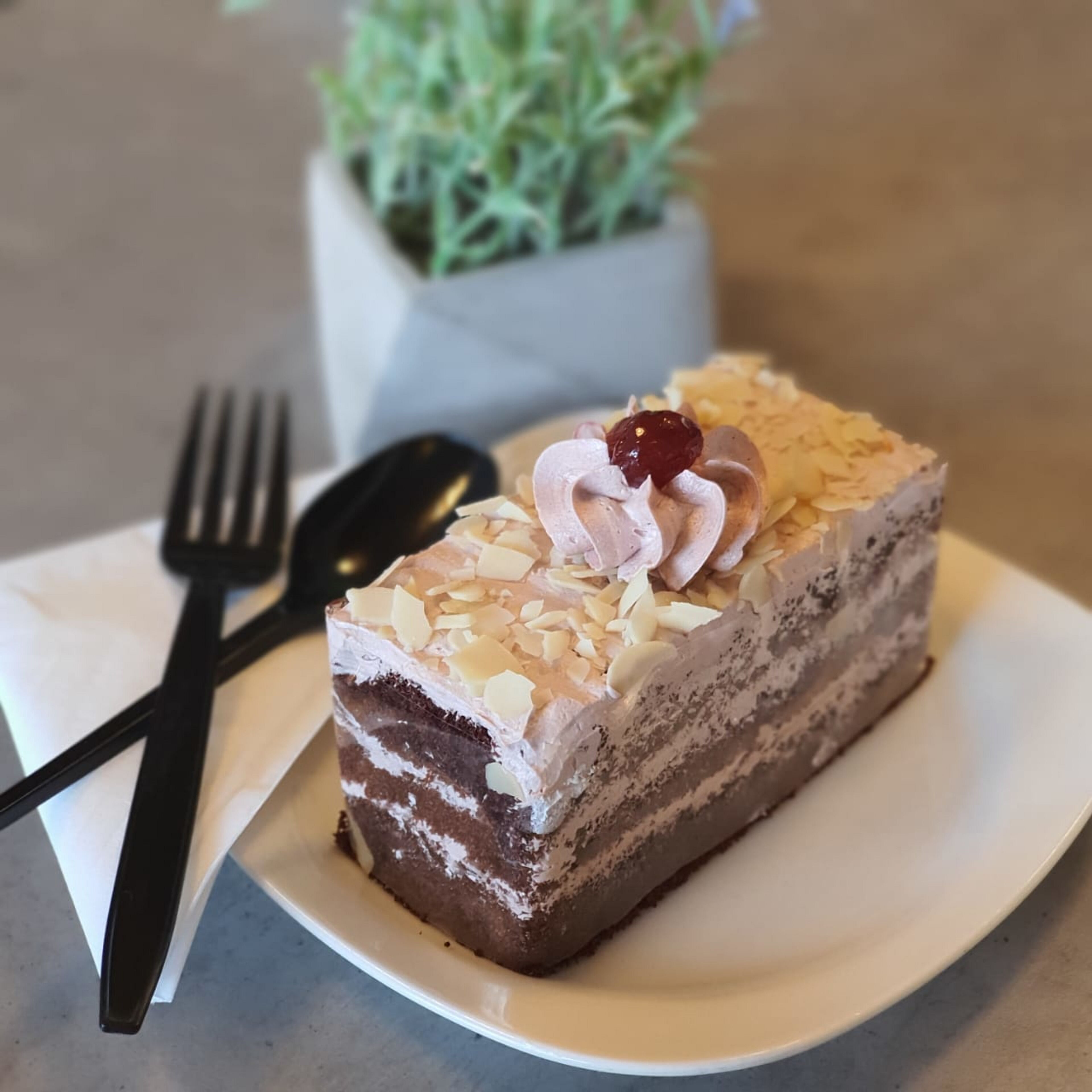 Une part de gâteau à plusieurs couches, aux amandes, surmontée d’une cerise, servie sur une assiette blanche avec une fourchette et une cuillère à côté.