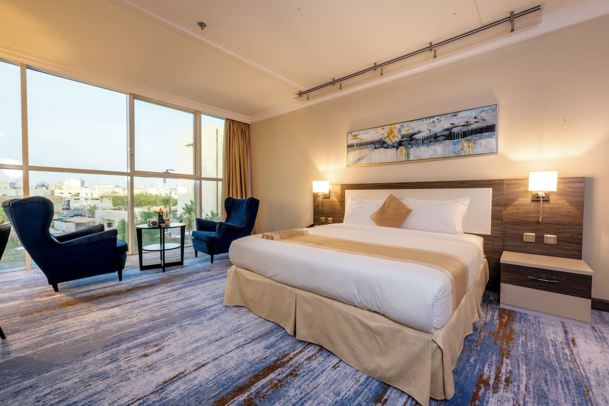 Habitación de hotel con una cama grande, dos sillones, una mesita y vistas a la ciudad a través de amplios ventanales.