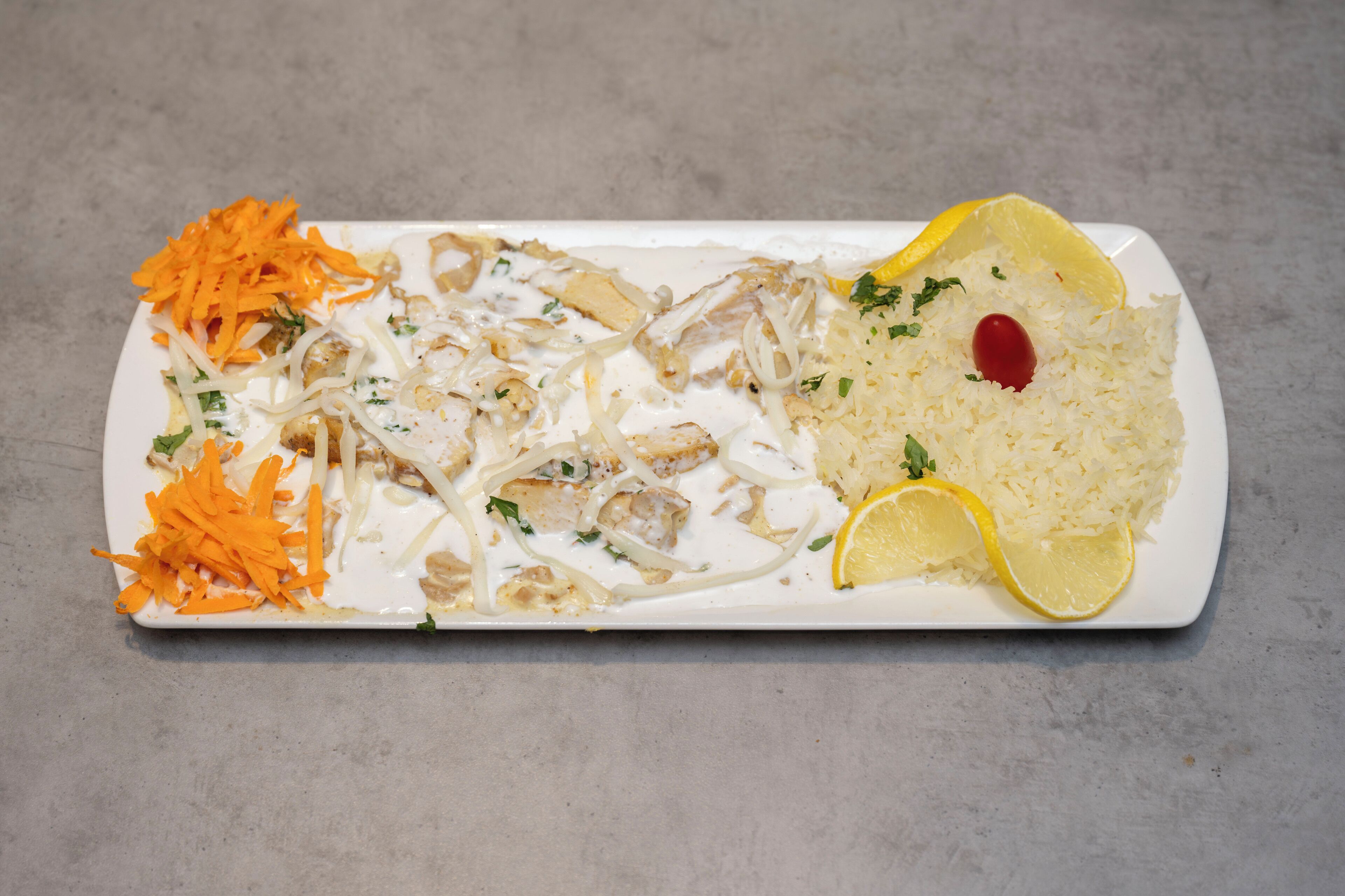 A rectangular white plate with shredded carrots, shredded chicken, and rice garnished with lemon slices and a cherry tomato.