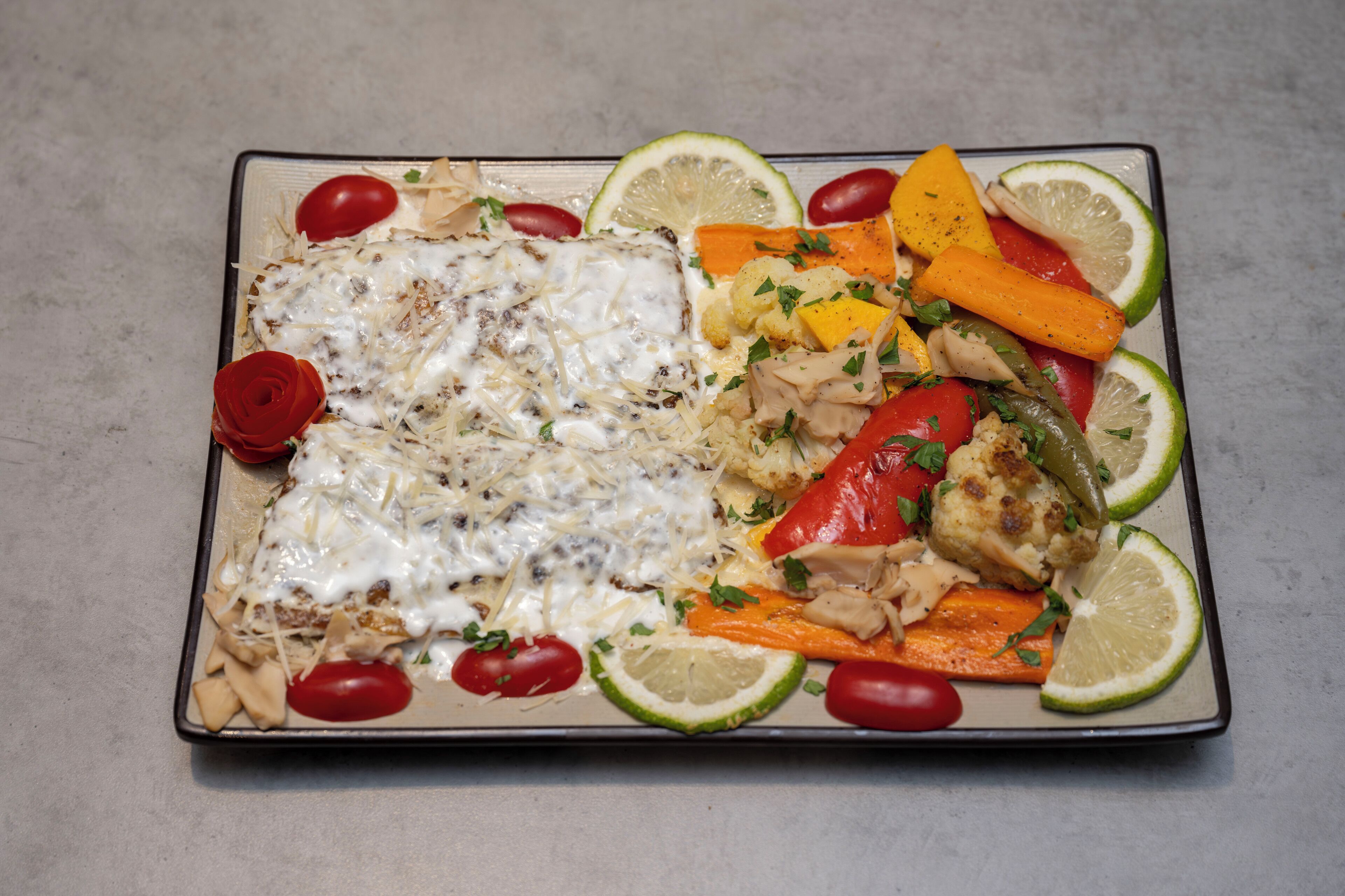 En rektangulär tallrik med ett urval av olika maträtter, inklusive en bit fisk täckt med sås, grillade grönsaker och garnerad med örter.