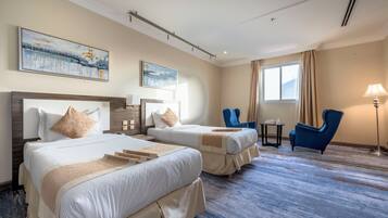 Quarto de hotel com duas camas, uma cadeira azul, uma mesa pequena e obras de arte nas paredes.