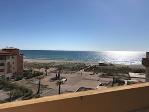 Udsigt over kysten med en strand, klar himmel og en bygning med balkon der kigger ud over havet.