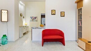 Una habitación con una silla roja, un escritorio con lámpara, una estantería y una pared con cuadros enmarcados.