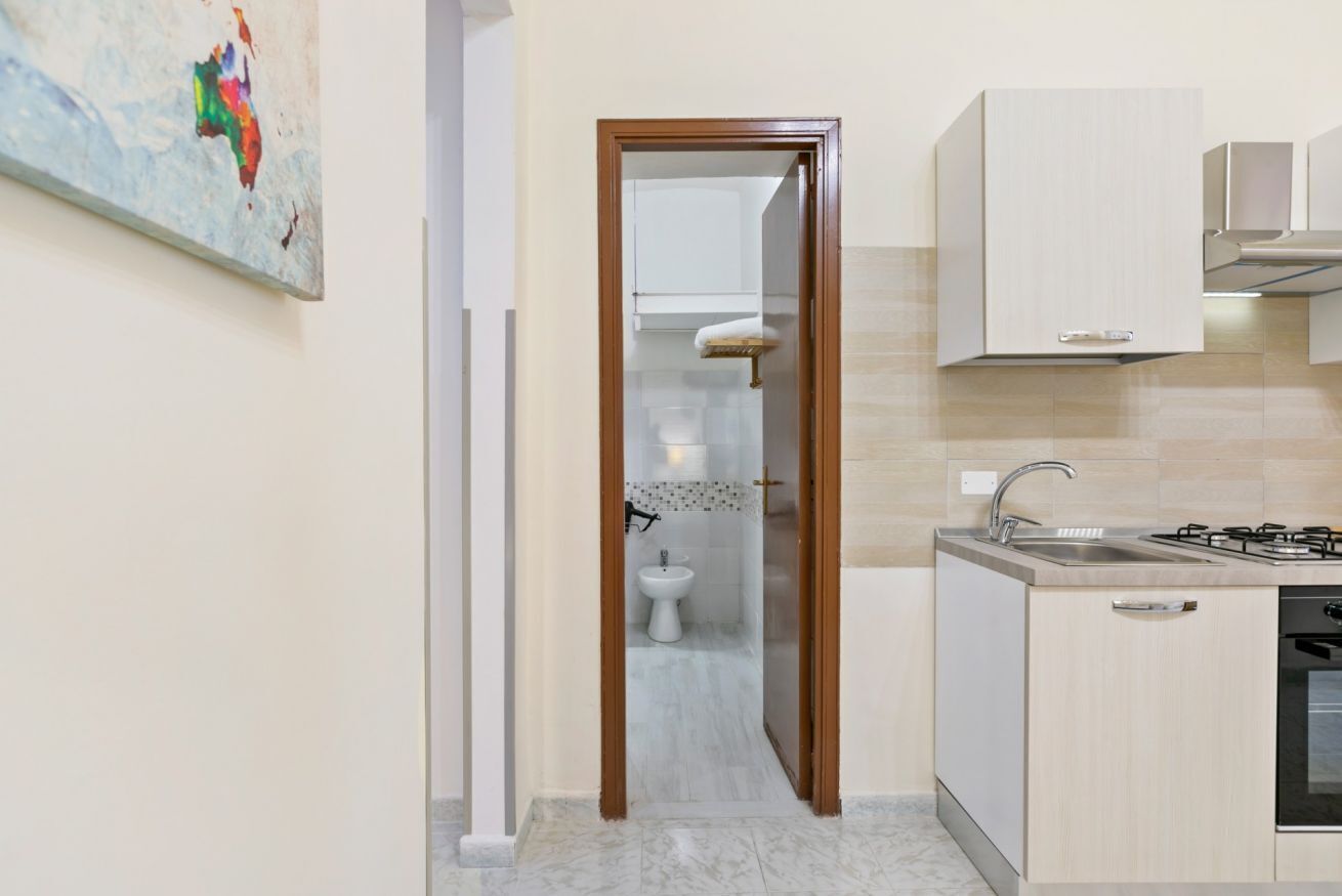Een compacte keuken met een spoelbak, kookplaat en kastjes. Een kleine badkamer is zichtbaar door een deuropening.