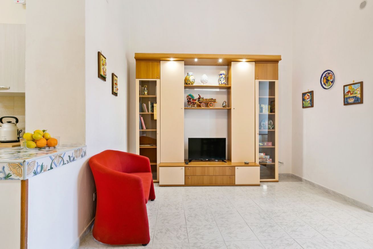 A living room with a TV, a red chair, and a kitchen area with fruit and a kettle.