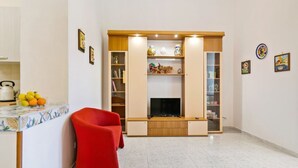 Una sala de estar con televisor, un sillón rojo y una zona de cocina con fruta y hervidor.