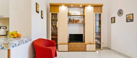 A living room with a TV, a red chair, and a kitchen area with fruit and a kettle.