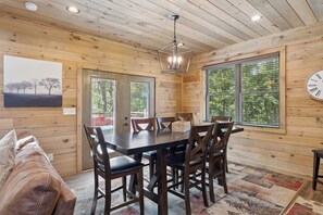 Interior de una cabaña de madera con una mesa de comedor, sillas, un sofá y un amplio ventanal con persianas.