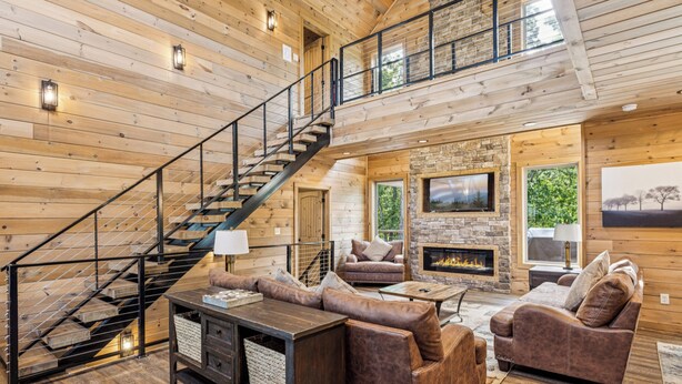 A spacious living area with a wooden interior, a stone fireplace, a television mounted on the wall, and a staircase with a metal railing. - Luxury 4 Bedroom with King Bedroom Suites, Hot Tub, Theater, WiFi, Game Room! (Sevierville)