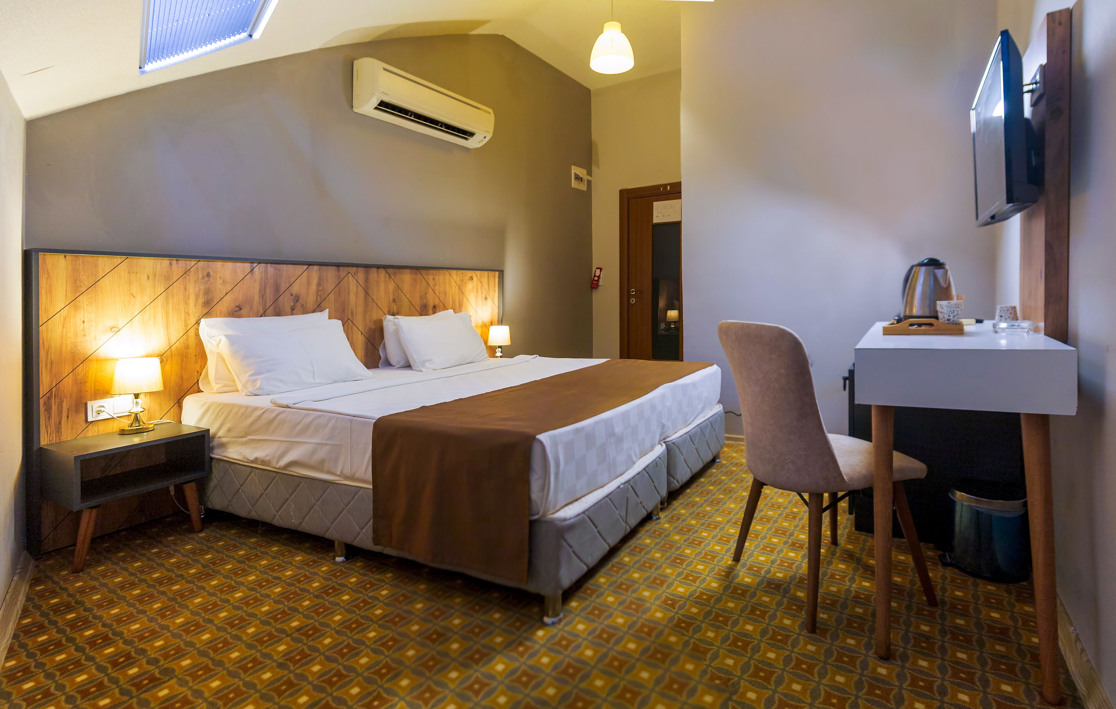 Una habitación de hotel con cama, mesitas de noche, escritorio, silla, equipo de aire acondicionado, televisor y piso con estampado.