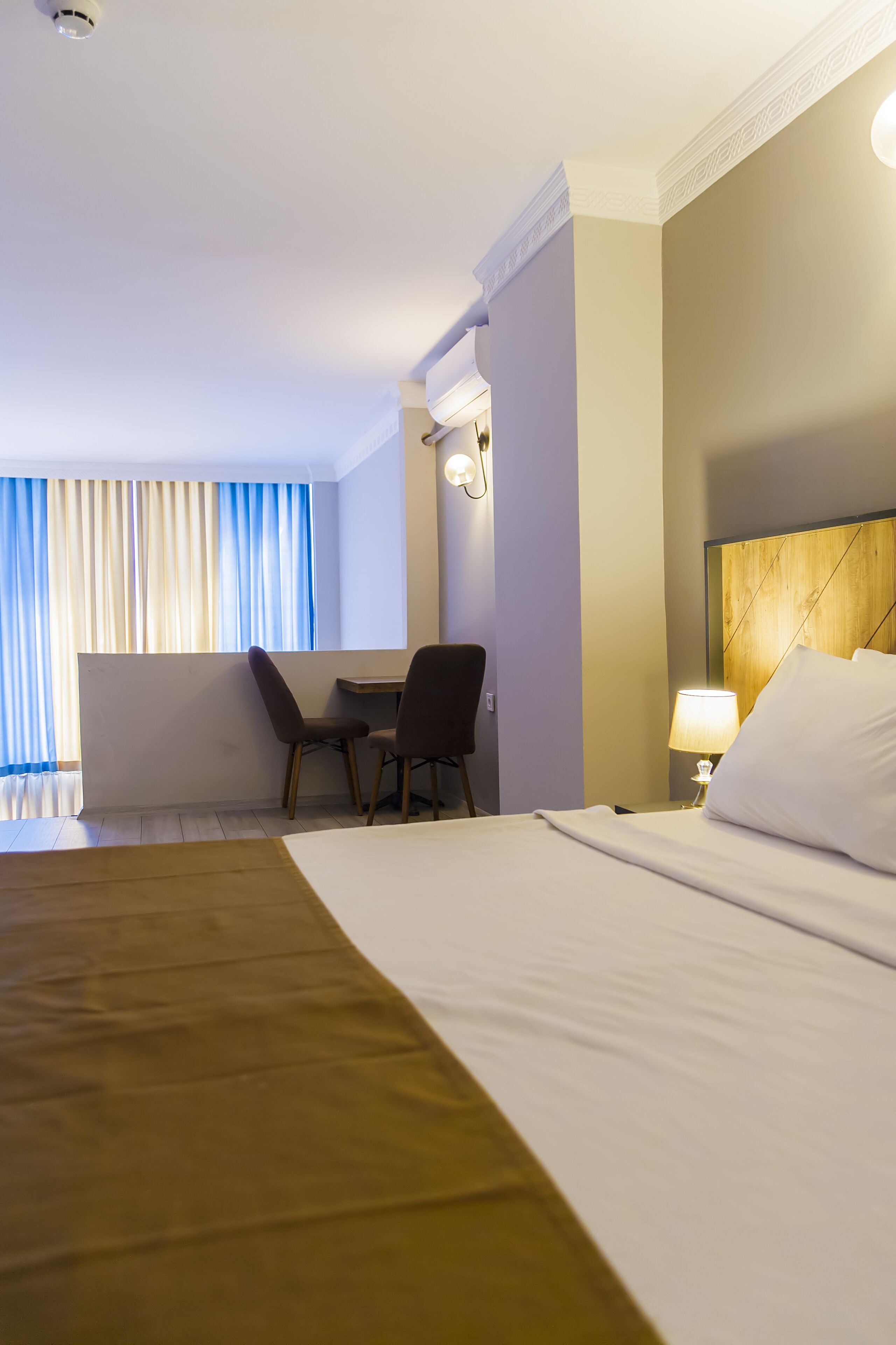 Habitación de hotel con una cama grande, un escritorio con dos sillas, una lámpara y vistas al exterior a través de cortinas.