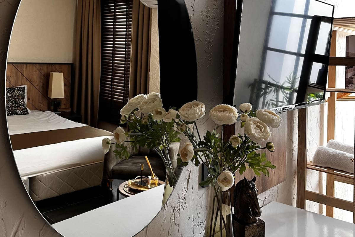 A hotel room with a large round mirror, a bed, a bedside table with a vase of flowers, and a flat-screen TV mounted on the wall.