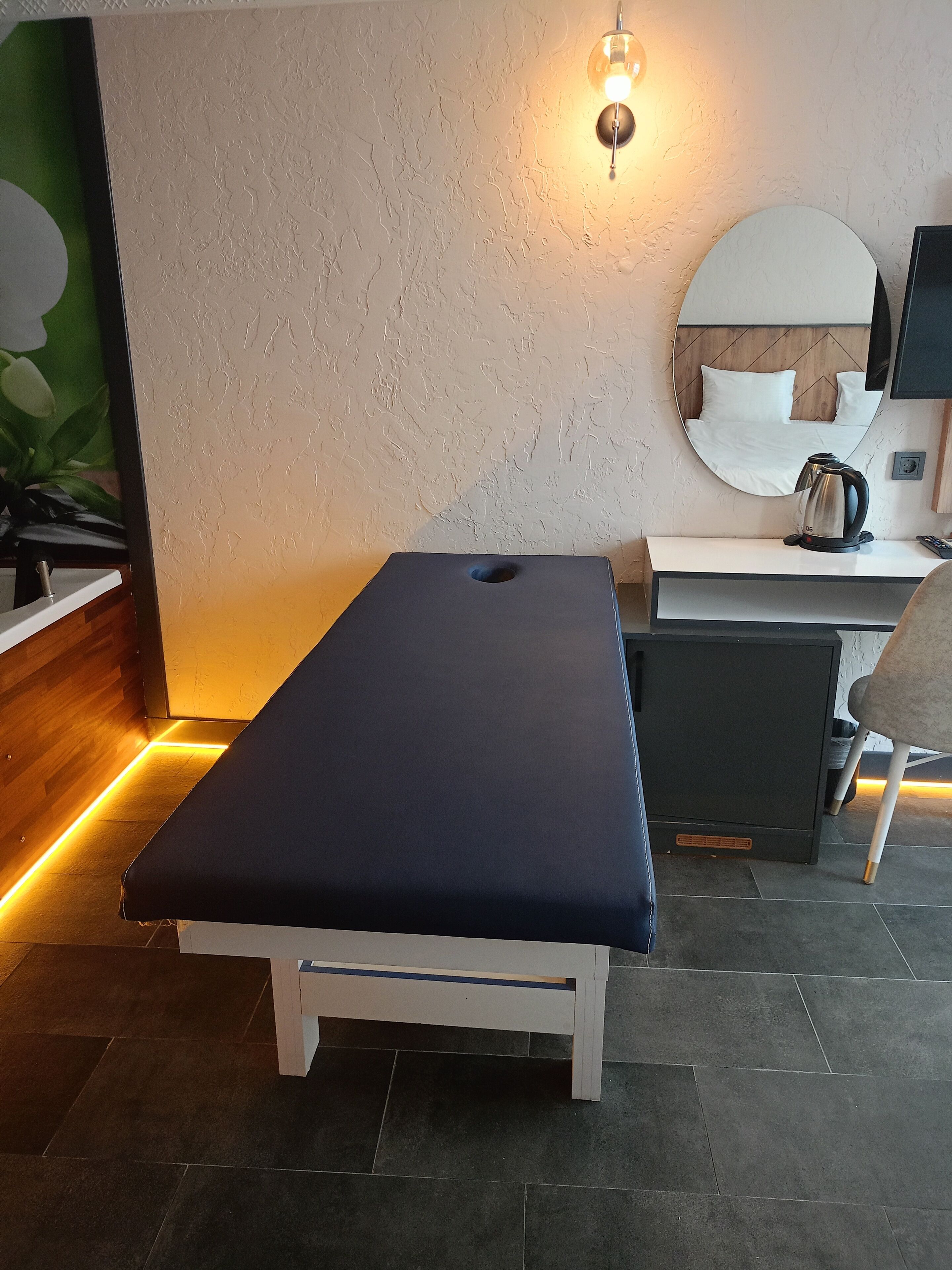 A massage table in a room with a wooden floor, a white wall with a textured finish, a round mirror, a small white desk with a kettle, and a chair.