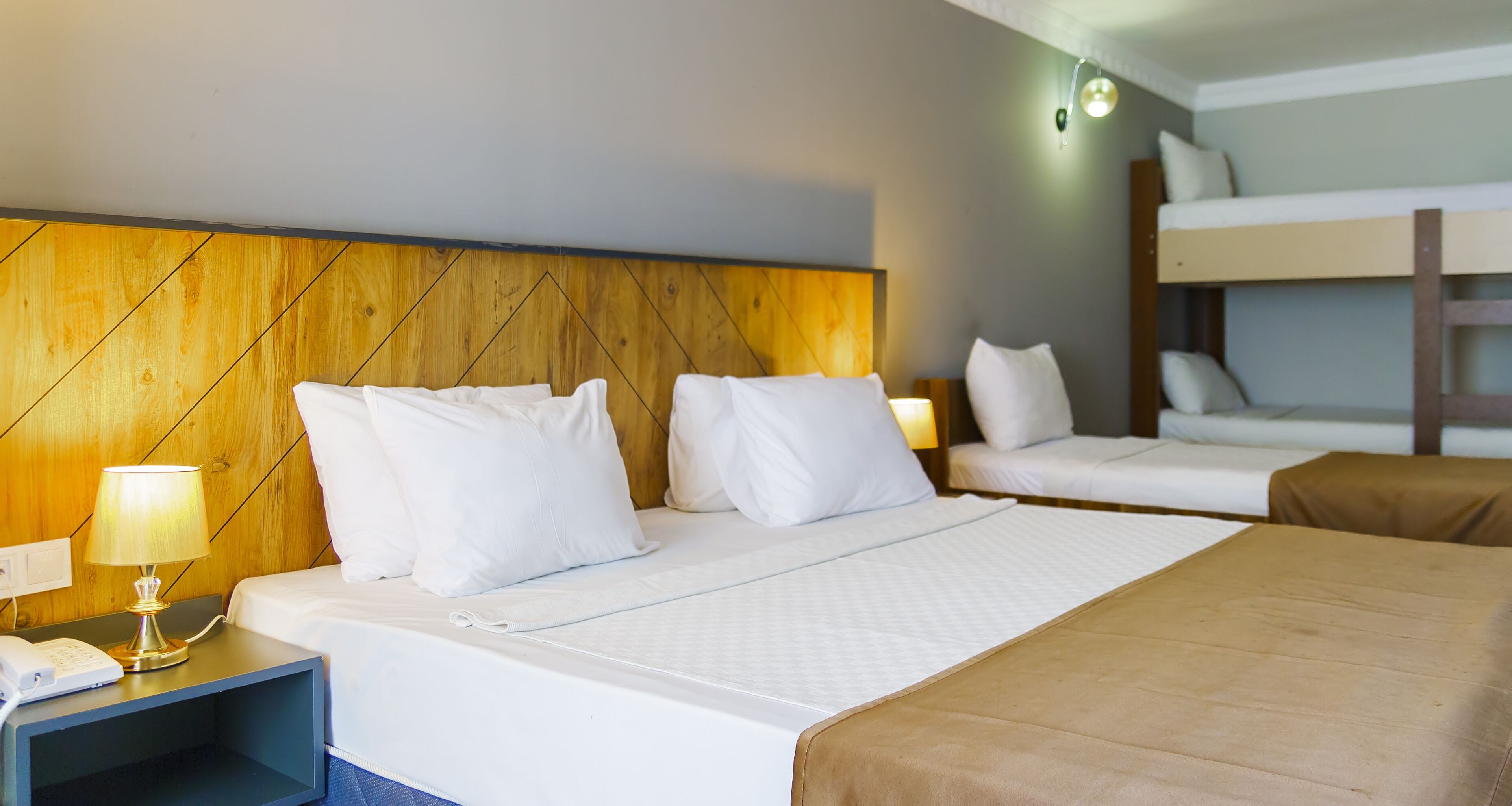 Habitación de hotel con cabecera de madera, cama con ropa blanca, lámpara de noche, teléfono y una cama litera al fondo.