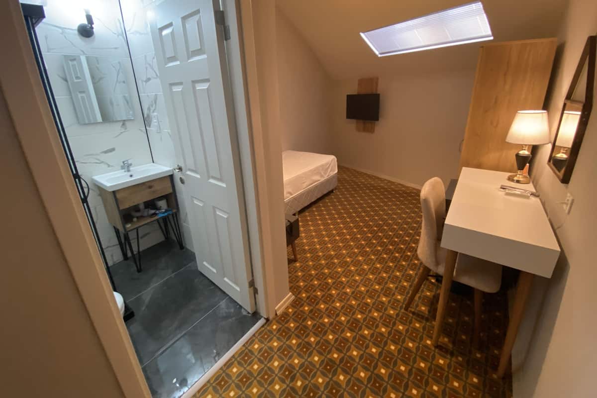 A compact bedroom with a bed, a bedside table with a lamp, a small desk with a chair, and a bathroom with a sink and mirror.