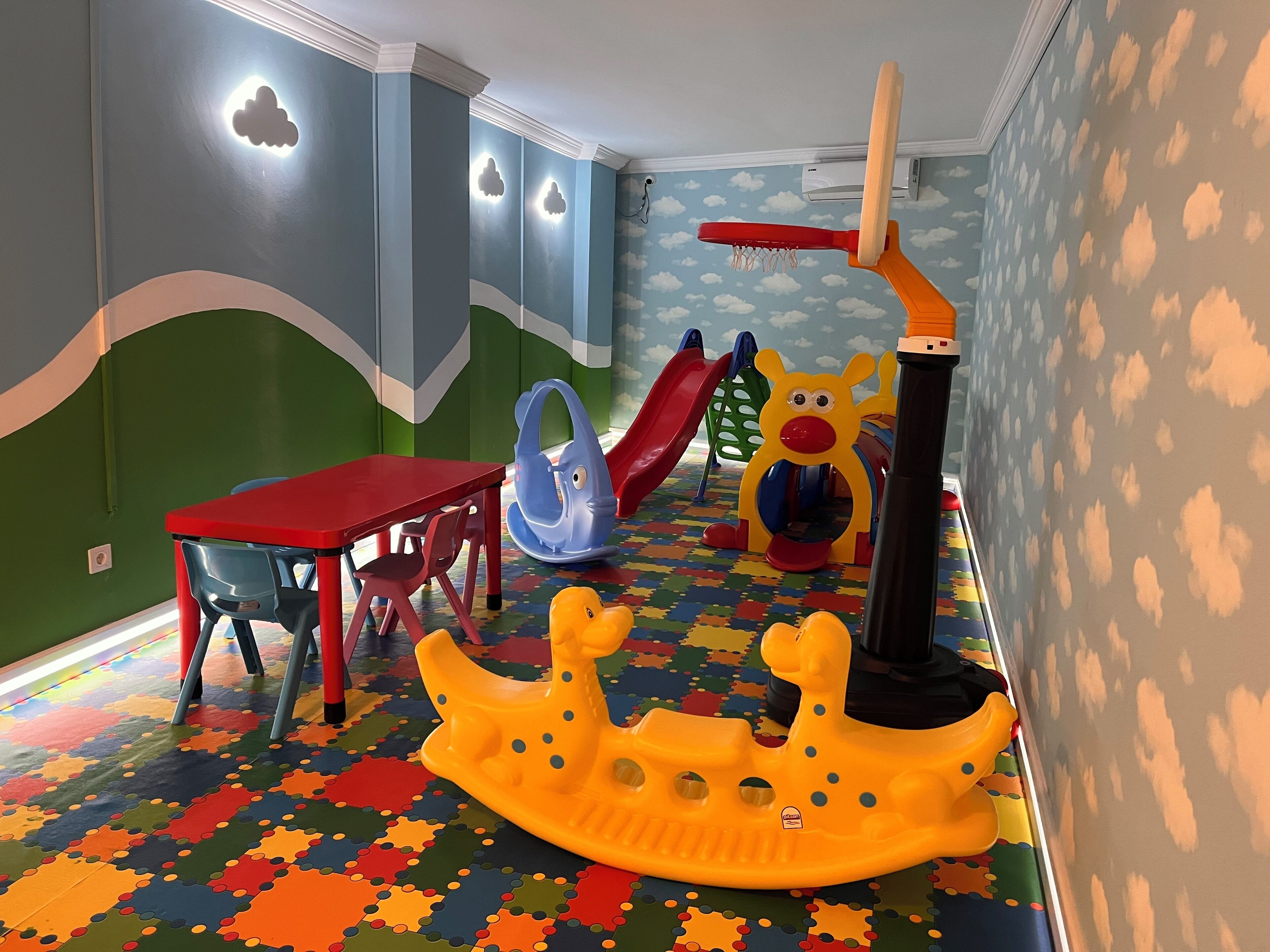 A playroom with a colorful floor, a red table, and various play structures including a slide and a climbing frame.