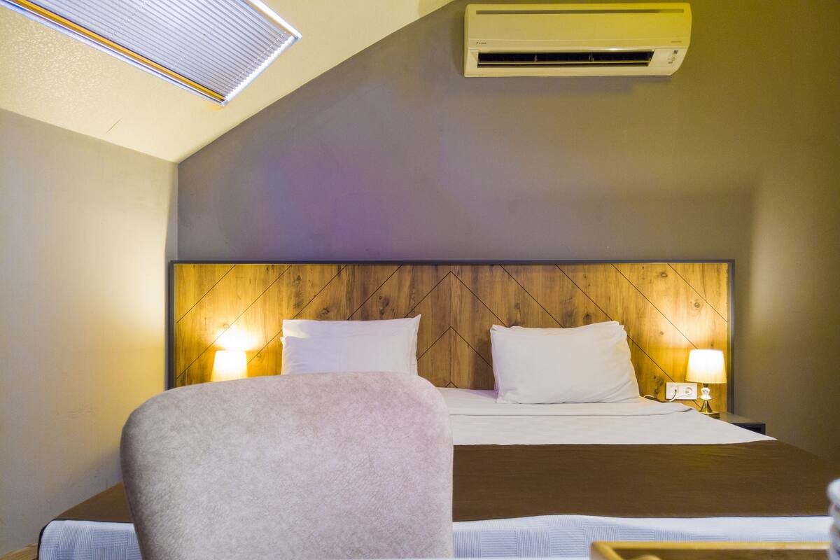 A hotel room with a bed, two bedside lamps, a wooden headboard, and an air conditioning unit.
