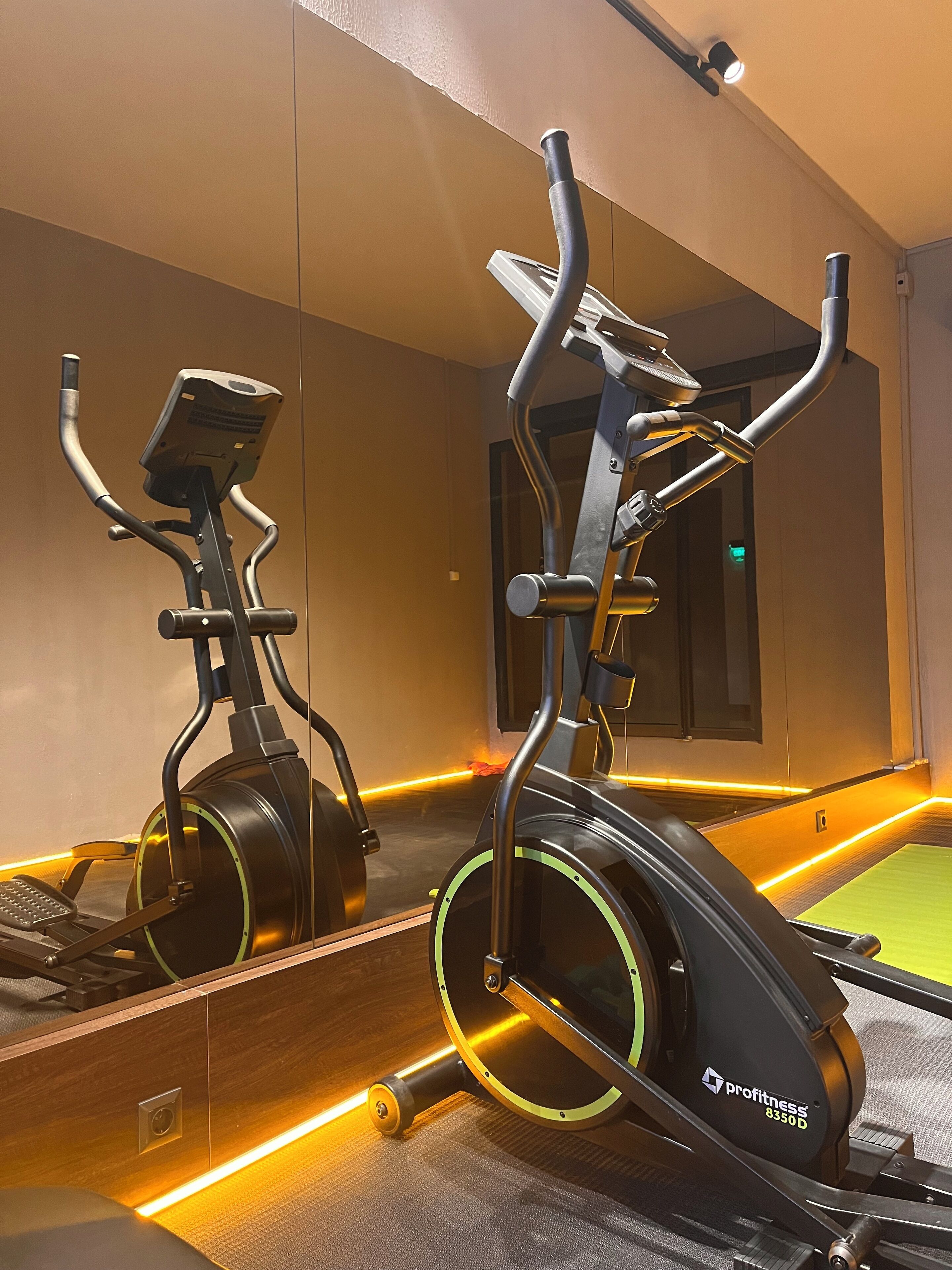 A stationary bike in a gym with a mirror reflecting the equipment and a green exercise mat.