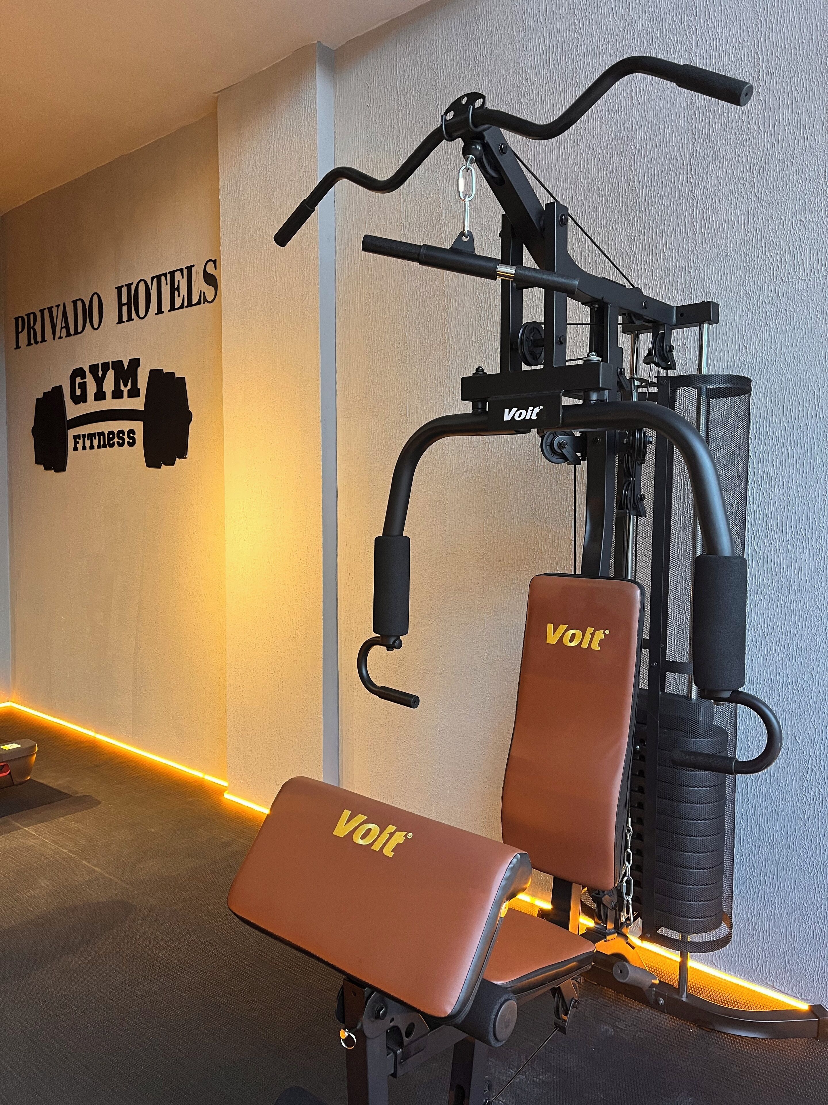 A gym with a wall sign reading "PRIVADO HOTELS GYM FITNESS" and a piece of exercise equipment branded "Volt".