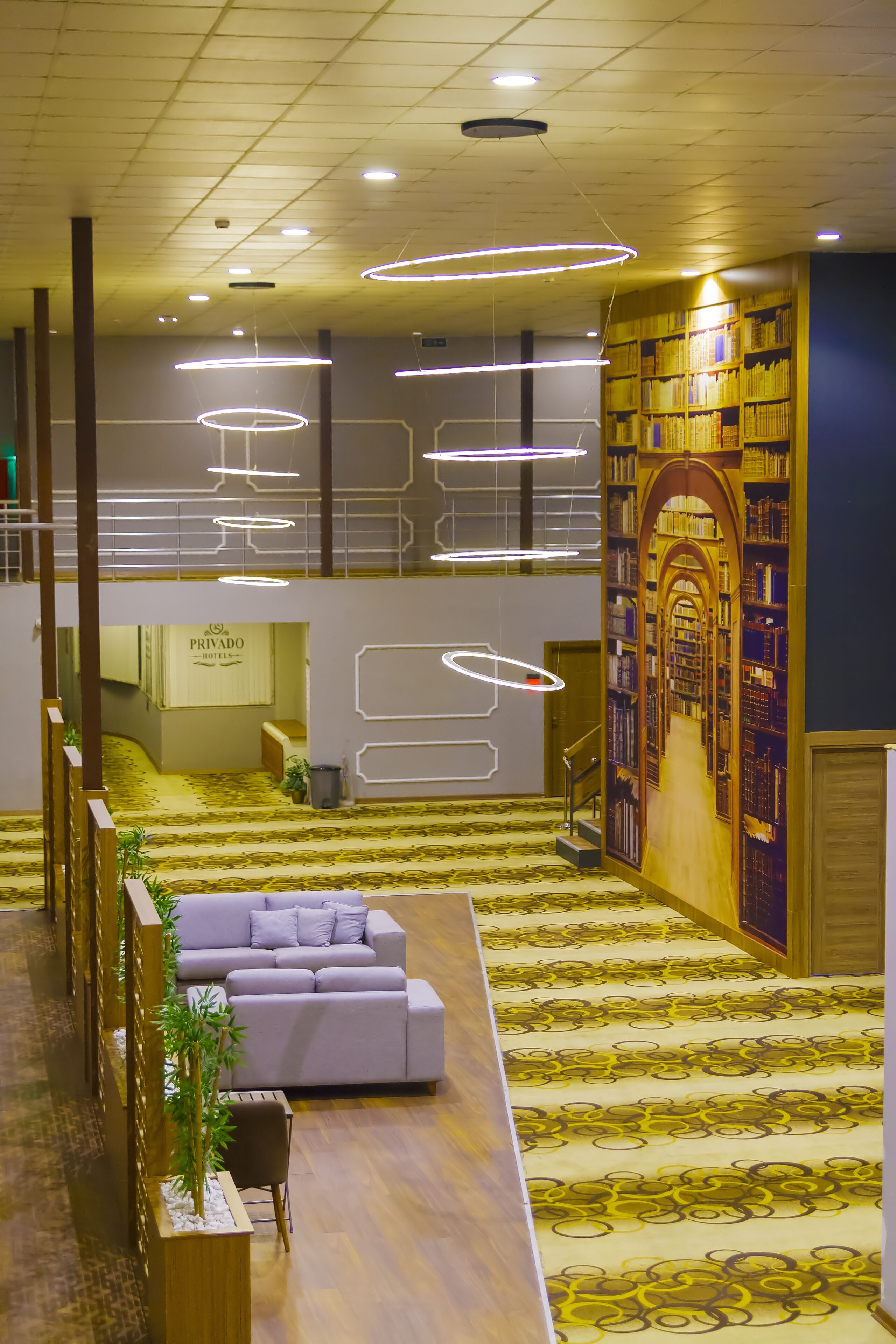 A hotel lobby with a modern design, featuring a central seating area, a staircase, and a large bookshelf.
