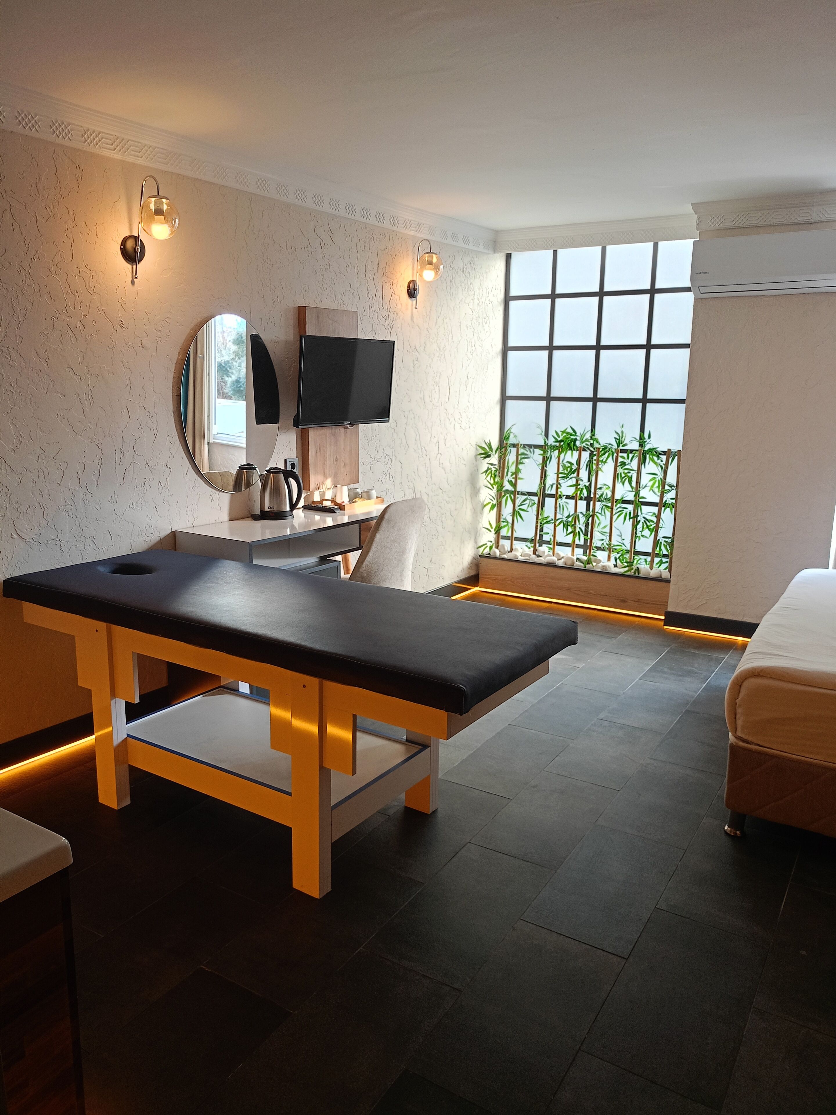 A modern room with a massage table, a television, and a large window with a view of bamboo plants.