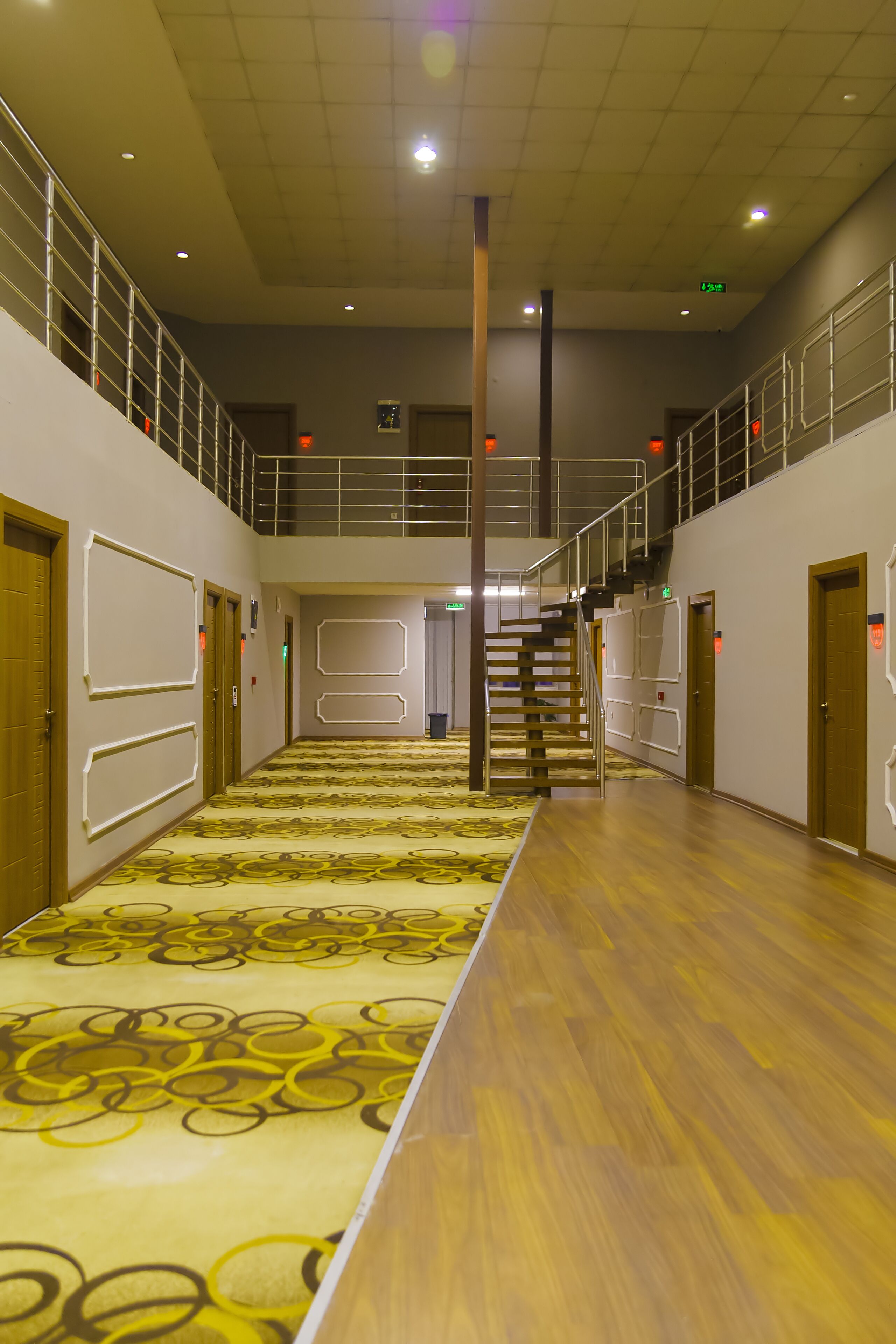 A spacious hotel corridor with a wooden floor, a central staircase, and a mezzanine level with a railing.