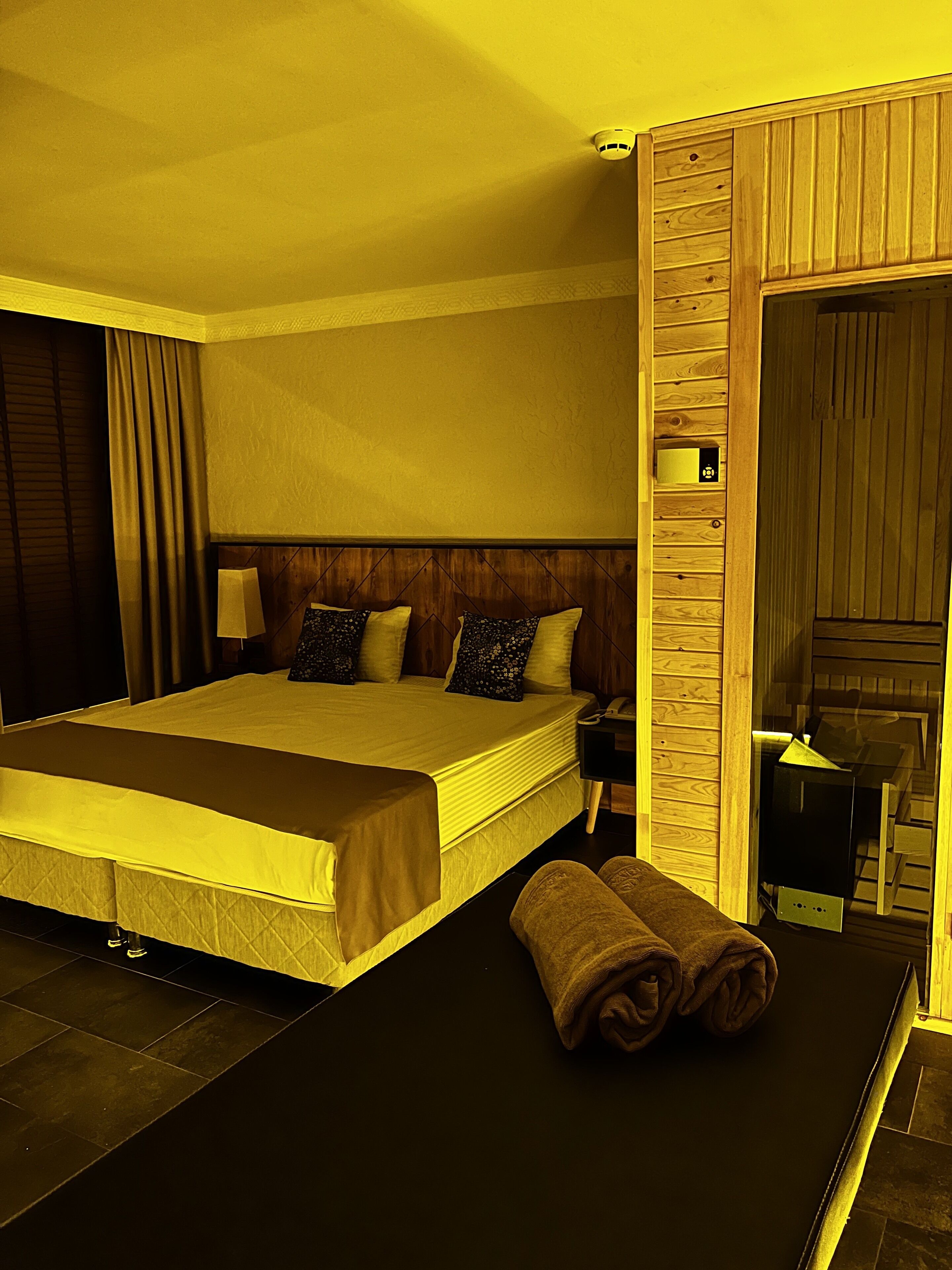 A hotel room with a large bed, wooden headboard, a bedside table with a lamp, and a bathroom visible through an open door.