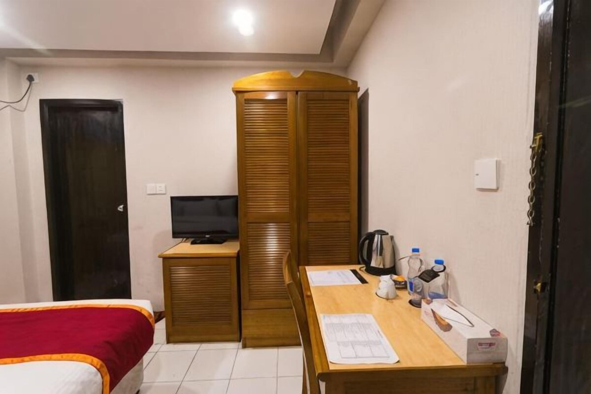 A bedroom with a bed, a wooden wardrobe, a desk with papers, a kettle, and a television.
