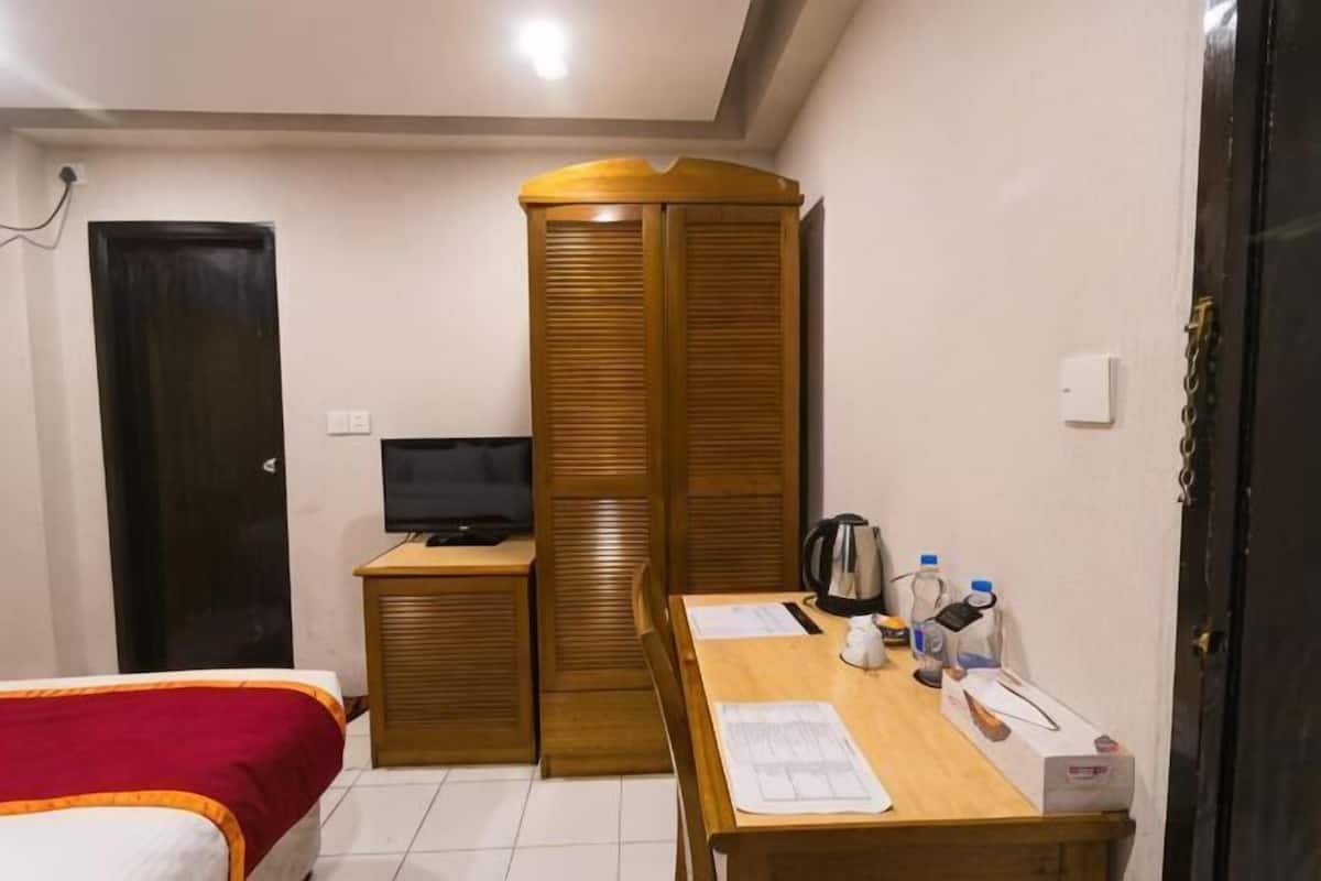 A bedroom with a bed, a wooden wardrobe, a desk with papers, a kettle, and a television.