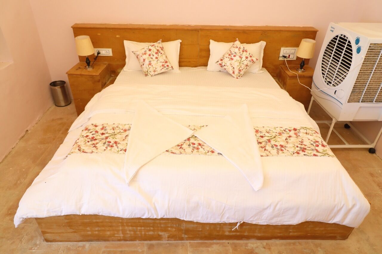 A neatly made bed with floral-patterned pillows and a white bedspread. There is a bedside table with a lamp and a small trash can. An air conditioning unit is visible to the right of the bed.