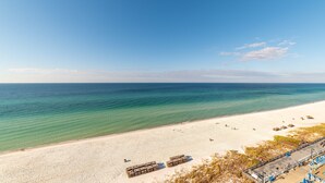 Une plage avec une eau turquoise et limpide, du sable blanc et quelques personnes qui se prélassent au soleil. On y trouve des structures en bois et un espace abrité avec des chaises bleues.