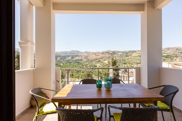 A balcony with a wooden table and wicker chairs, overlooking a mountainous landscape.