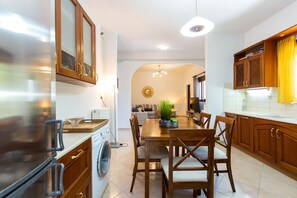 A modern kitchen with wooden cabinets, a built-in washing machine, a dining area with a table and chairs, and a view into the living room.