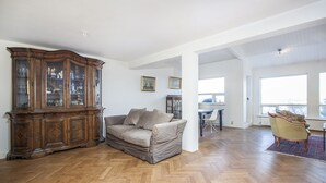 En rummelig stue med parketgulv i fiskebensmønster, et stort træskab, en beige sofa og et spiseområde med bord og stole.
