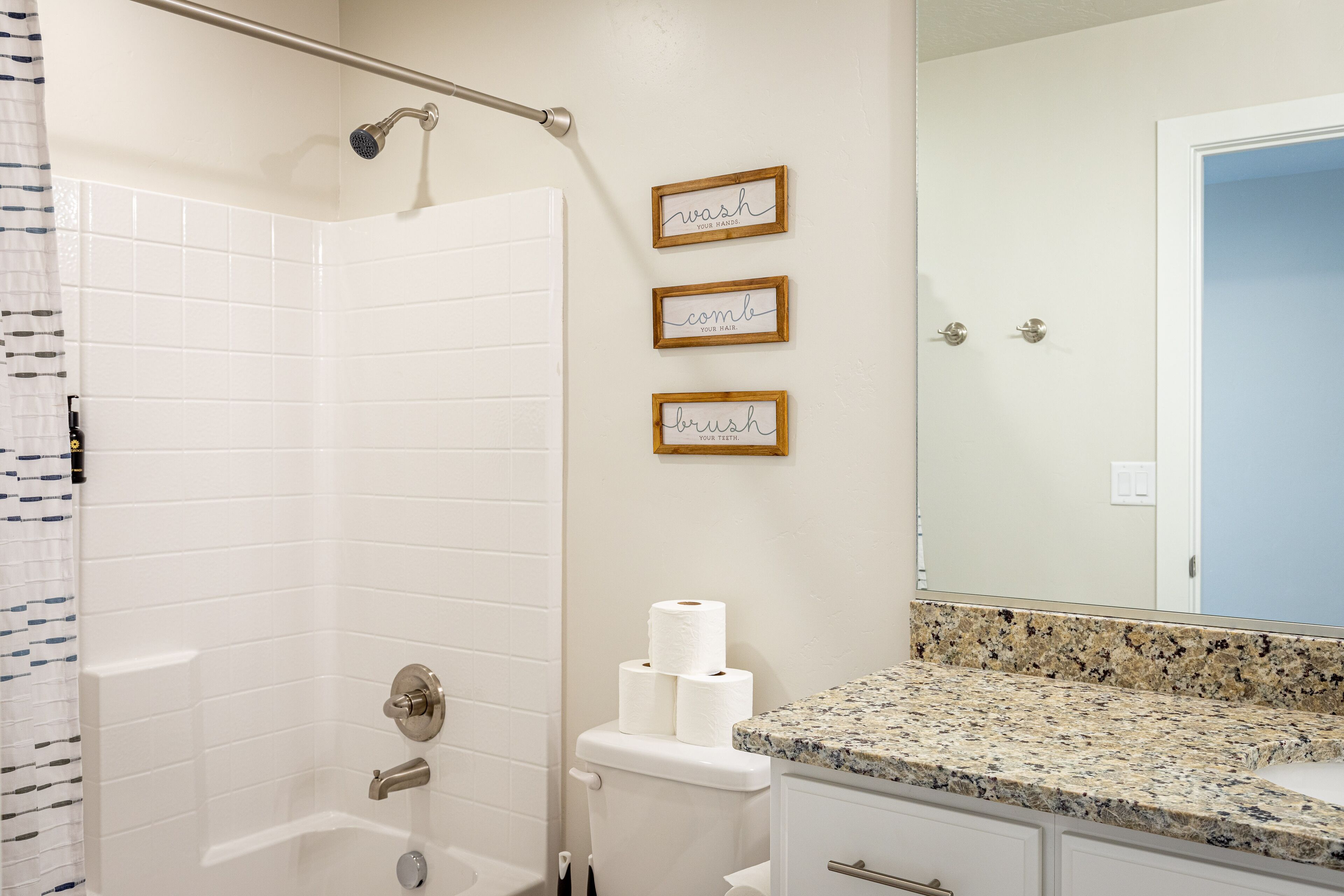 A bathroom with a shower, toilet, and vanity with a mirror. There are three framed signs on the wall.