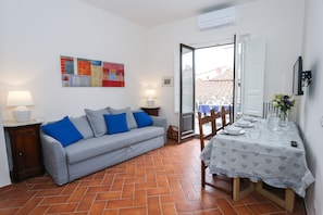 A living room with a grey sofa, blue cushions, a dining table with a white tablecloth, and a view through an open door to the outside. - gorgeous terrace in Florence (Firenze)