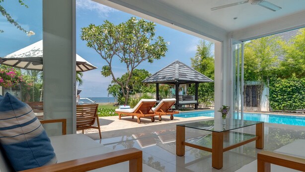 A modern outdoor lounge area with a glass table, wooden chairs, and a pool view.