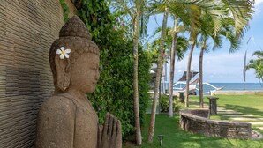 A Buddha statue with a white flower, surrounded by lush greenery and palm trees, with a view of the ocean and a building in the background.